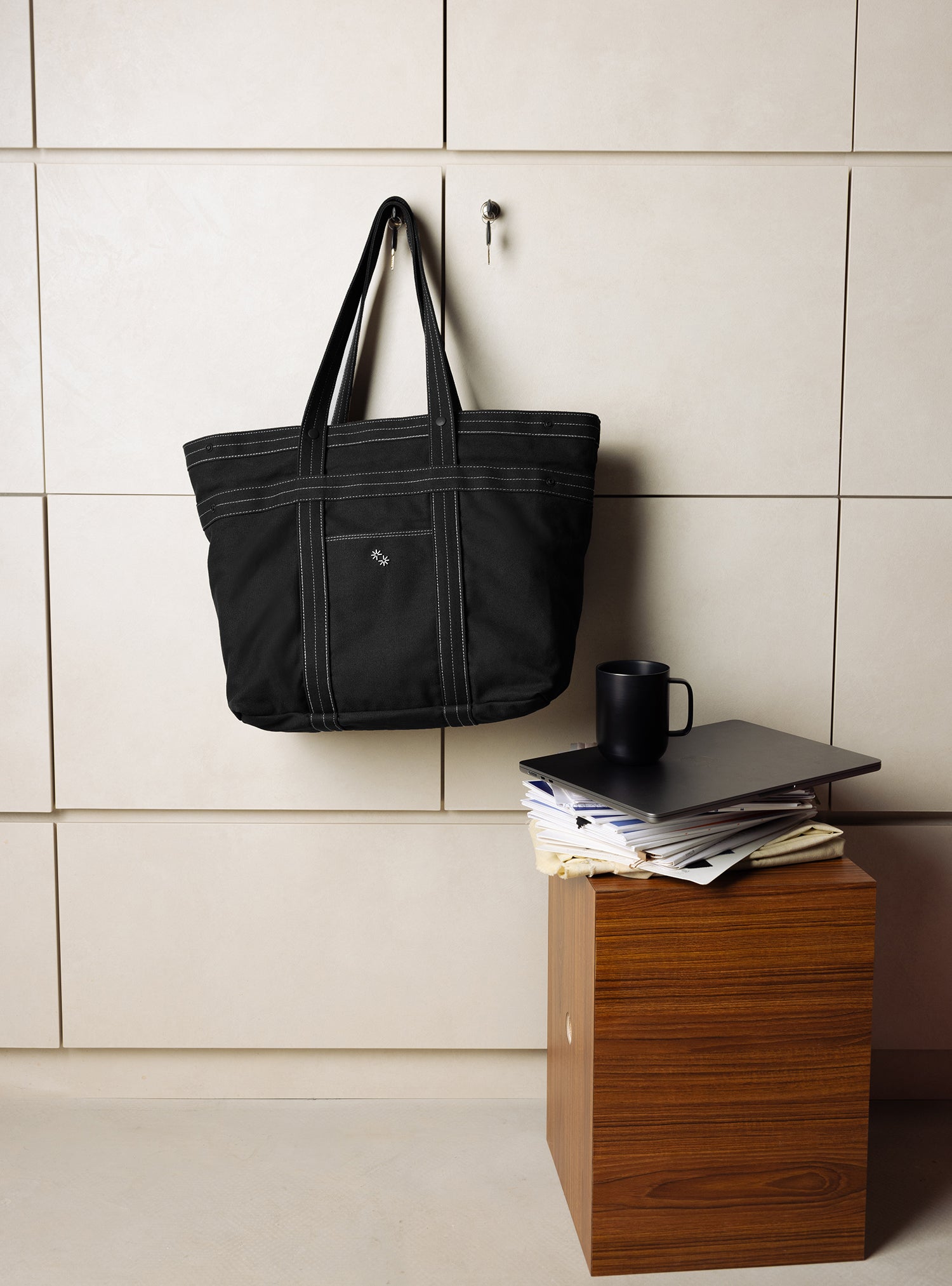 Black tote bag hanging on a tiled wall next to a wooden block with a coffee mug and books.